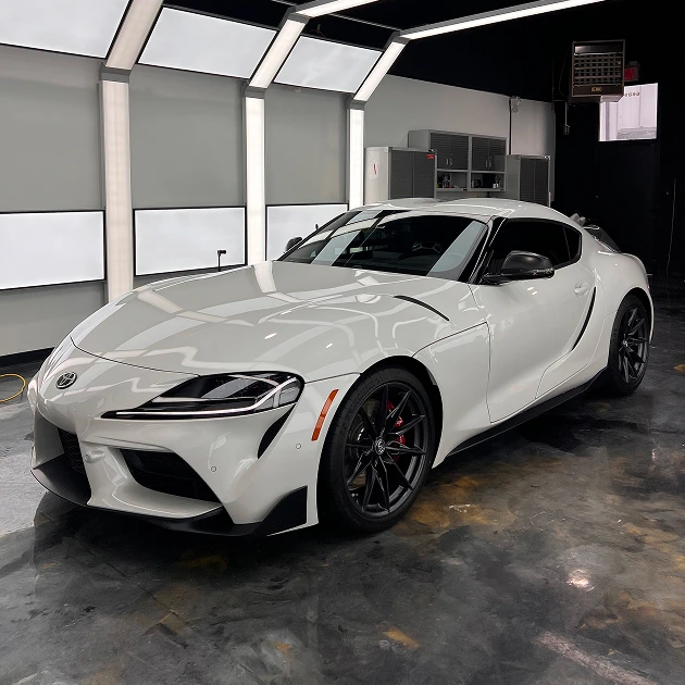 White Toyota Supra parked in a clean, modern auto detailing bay with reflective black flooring
