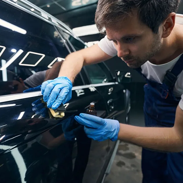 Man in blue gloves and overalls applying ceramic coating to the side of a black car with a sponge and a small bottle