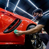 Auto detailing technician in a face mask polishing the side of a red sports car under LED ceiling lights