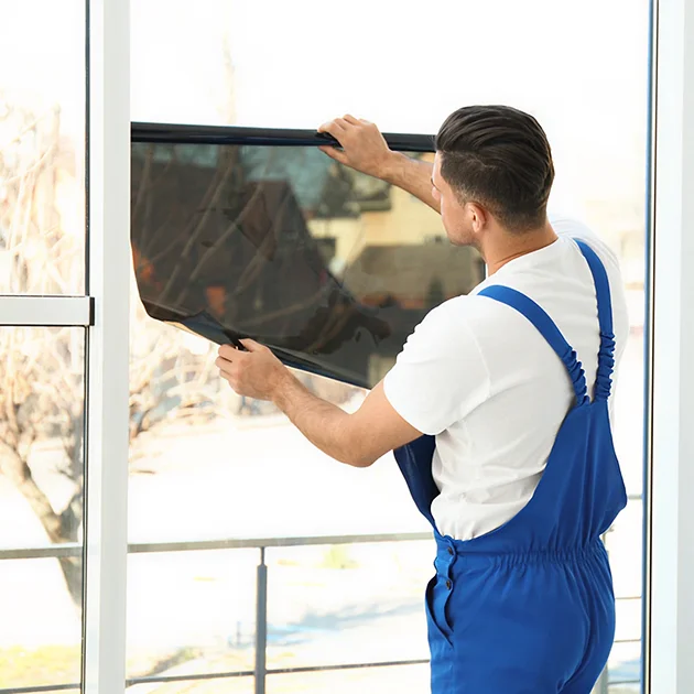 Technician in blue overalls applying dark tint film to a large glass window in a bright residential space