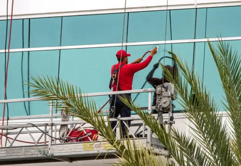 Window cleaner in red uniform using a squeegee on the upper floors of a glass building while standing on a suspended platform
