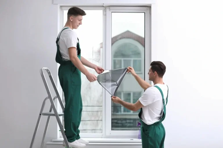 Two technicians in green overalls applying tint film to a residential window, one on a ladder and one holding the film from below