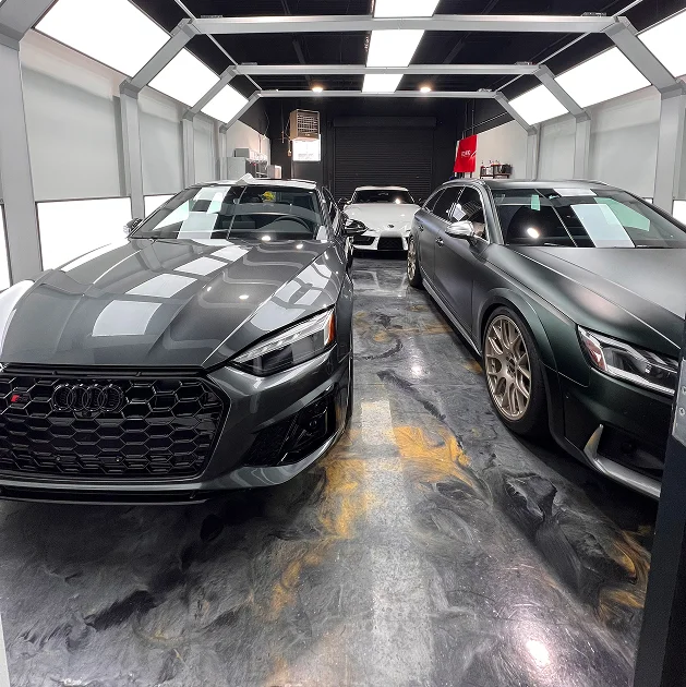Three luxury cars inside a detailing bay under bright overhead lights, including two dark Audi models and a white sports car in the back