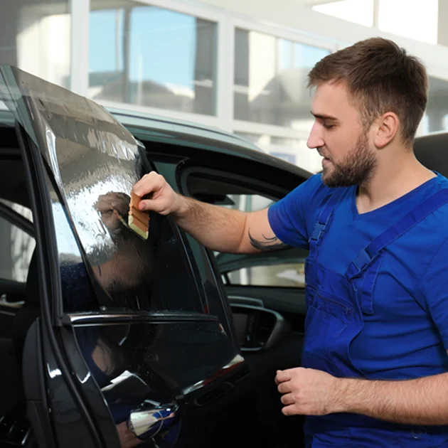 Auto technician in blue overalls applying tint film to a car window using a smoothing tool