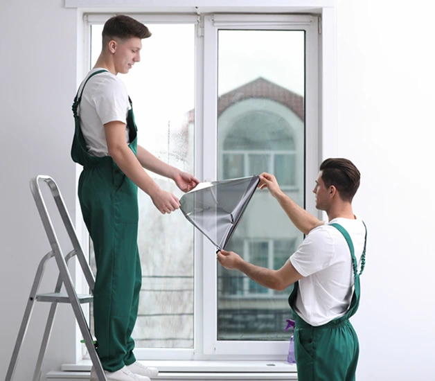 Two technicians in green overalls applying tint film to a large residential window, one standing on a step ladder